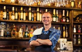 Smiling bartender stands before well-stocked bar ready to serve representing Award for Licensed Premises Staff Online course.