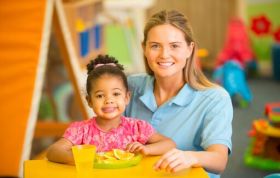 Smiling woman with child and kids playing behind, representing CACHE Level 3 Award in Childcare and Education course.