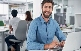 Bearded man in blue shirt working on laptop at desk, representing the CompTIA Cloud Essentials course for foundational cloud knowledge.