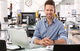 Man with a friendly smile sits at a desk, working on a laptop, representing the CompTIA IT Fundamentals course.