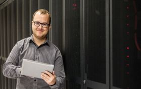 Man wearing glasses holding tablet in front of server rack, representing the CompTIA Mobility+ course for mobile device and network management.