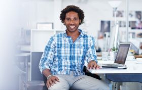Smiling man in checkered shirt in front of computer, representing the CompTIA Security+ (SYO-401) Course for cybersecurity fundamentals.