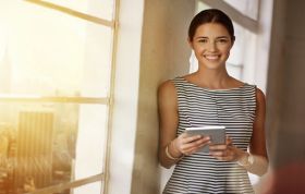 A woman stands by a window, holding a cell phone and looking thoughtfully, representing the Developing ASP.NET MVC Web Applications Online course.