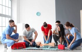 Man standing beside a "First Aid at Work" sign, indicating a safety area, representing the First Aid At Work Online course.