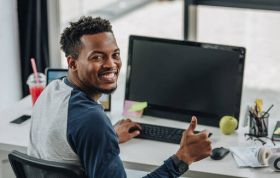A smiling man works on a computer at his desk, representing the Google DevOps Master Training Series course.