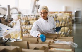 Woman in a white lab coat works in a factory with equipment, representing the Level 2 Food Safety for Manufacturing Online course.
