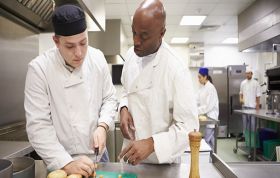 Two chefs in white uniforms prepare food in a commercial kitchen, representing the Level 3 Supervising Food Safety Online course.