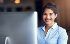 A cheerful woman sits in front of a computer, engaged in work, representing the Managing App and Desktop Solutions with Citrix XenApp and XenDesktop 7.6 Online course.