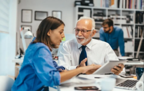 Smiling woman discussing with elderly man at desk, representing Managing People and managing tasks effectively in the workplace.