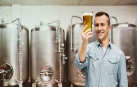 Smiling man raises a glass of beer in front of a brewery, representing the Mastering Beer Brewing course for craft brewing enthusiasts.