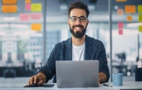 Smiling man sitting at desk with laptop, representing Microsoft Azure training for the AZ-104 certification exam preparation.