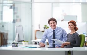 Man and woman at a desk discussing work with papers and a laptop, representing the Microsoft Excel 2010 Course in an office setting.