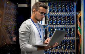 A man works on a laptop in a server room, representing the MTA Networking Fundamentals Online course.