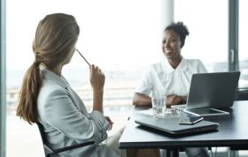 Two women talking at their desks with laptops, representing Performance Management and an effective appraisal system at work.