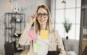 Woman with glasses holding tablet and looking at sticky note, representing the PRINCE2 Practitioner Online Course for project management skills.