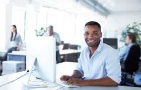 Cheerful man in front of a computer in a professional setting, representing the Complete CompTIA Bundle course for comprehensive IT training.