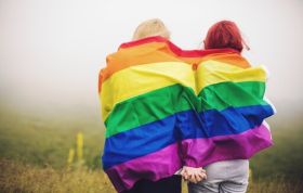 Two women holding hands with LGBT flag in background, representing LGBT Awareness and efforts to reduce stigmatism and promote inclusion.