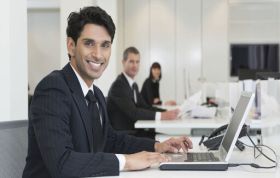 Man at laptop with two colleagues at desks, representing Windows 7 Enterprise Desktop Administrator Online Course.